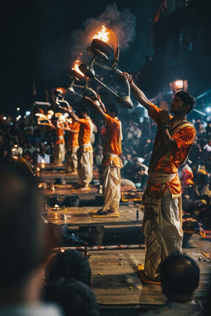 A group of Hindu priests in traditional orange robes holding large, flaming oil lamps during the Ganga Aarti ceremony at night in Varanasi, India.