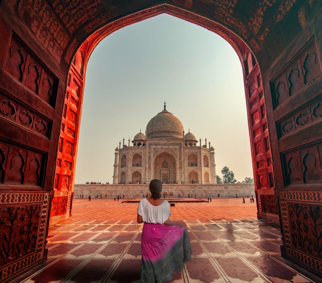 Top 10 Tourist Places in India; View of the Taj Mahal from inside a red sandstone archway, with a woman in the foreground looking out at the monument.