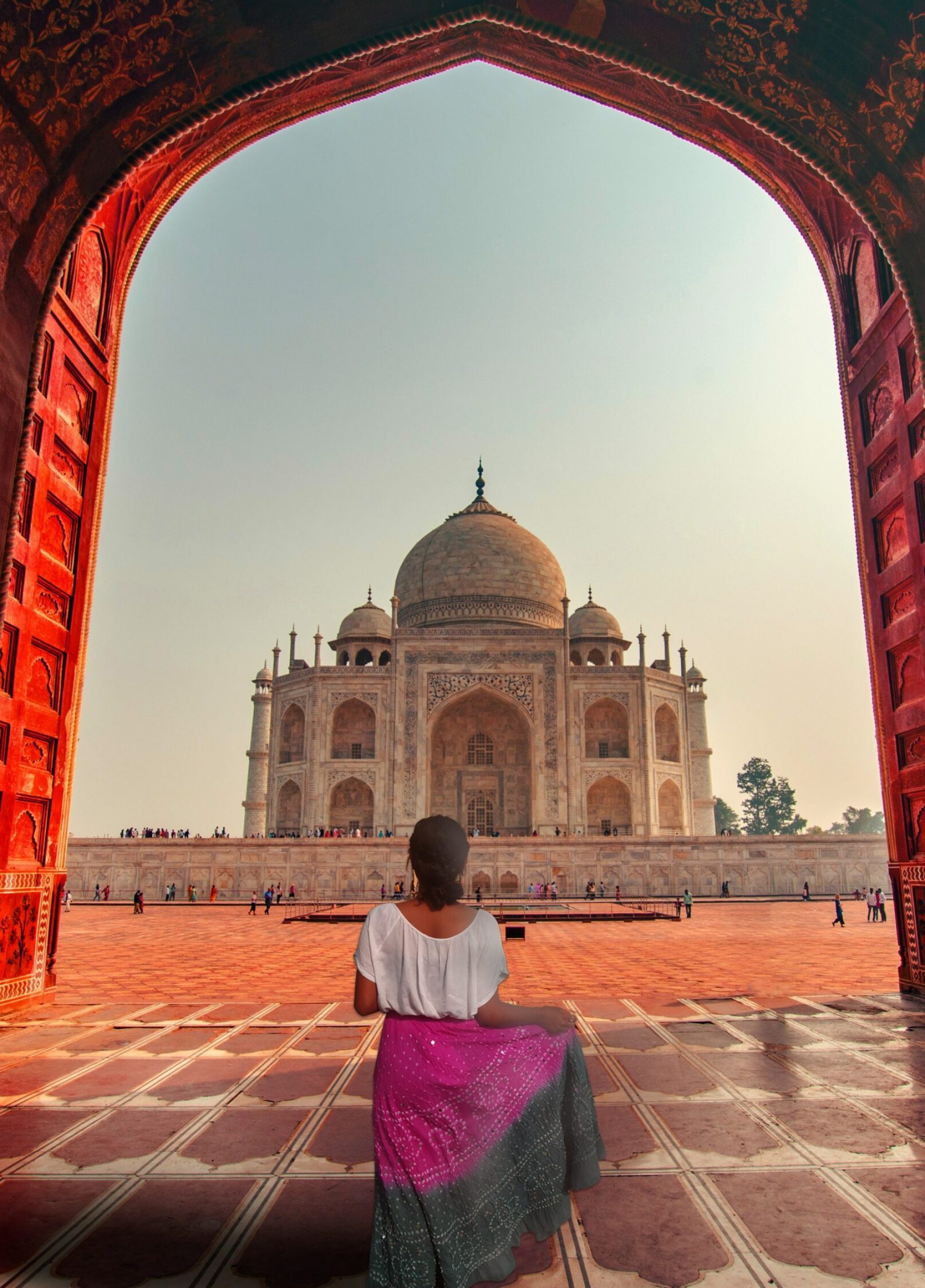 Top 10 Tourist Places in India; View of the Taj Mahal from inside a red sandstone archway, with a woman in the foreground looking out at the monument.
